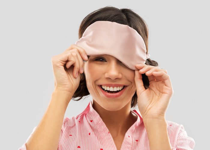 people and bedtime concept - happy young woman in pajama peeking from under sleeping mask over grey background